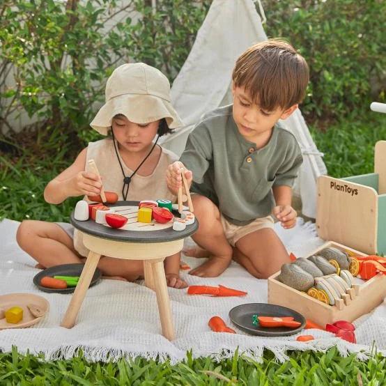 PlanToys wooden table BBQ playset with heart-shaped grill, utensils, and food accessories for imaginative pretend cooking.